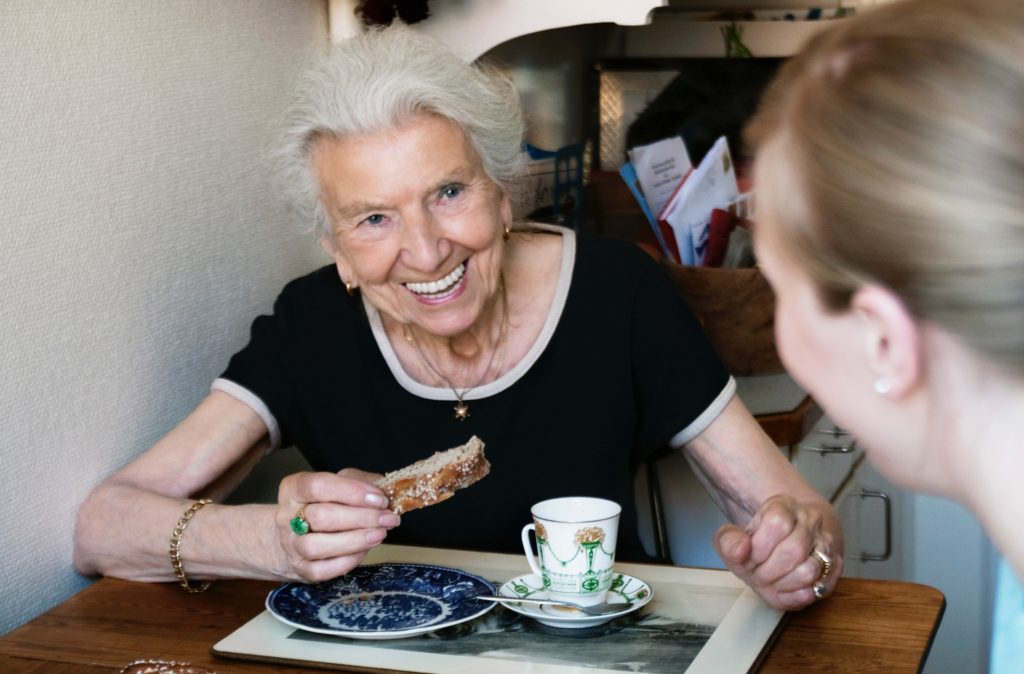 Une femme âgée souriante attablée à son domicile accompagnée d'une professionnelle de l'accompagnement à domicile